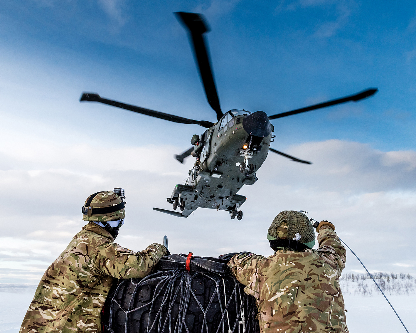 Image shows aviators tending to cargo load with helicopter taking off in snowy environment.